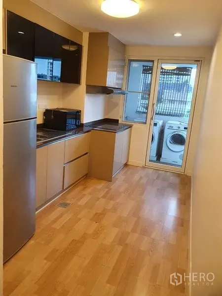 Condominium for rent in Watthana, Bangkok - Galley kitchen with modern cabinets, fridge, microwave, and glass door to a laundry balcony with washer and dryer.