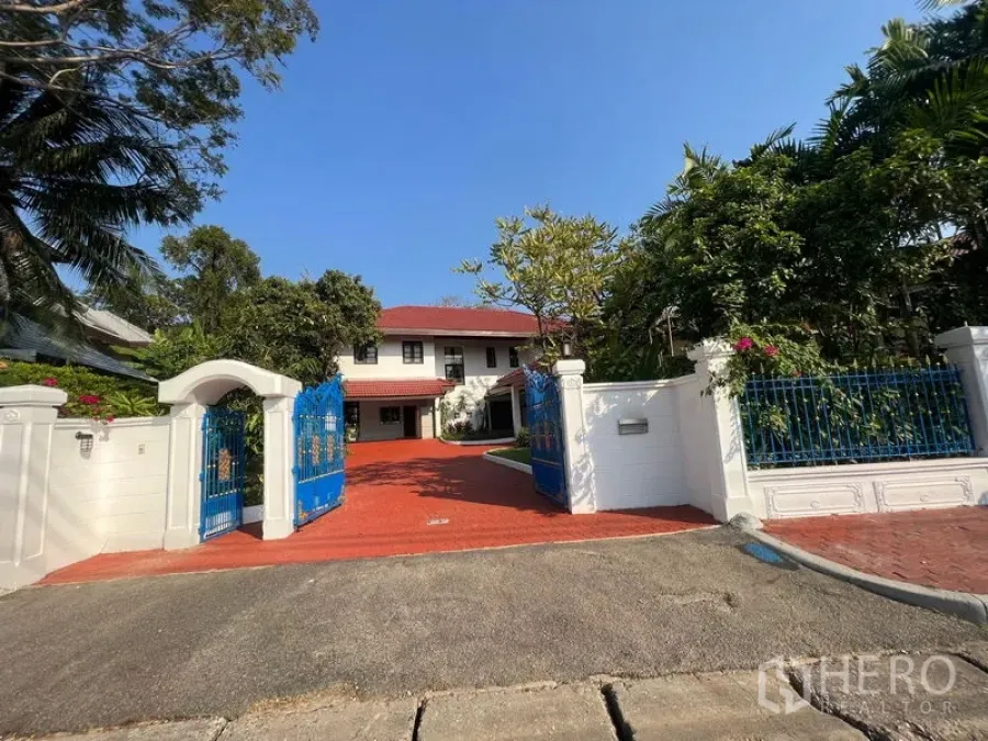 Detached House for rent in Bang Phli, Samut Prakan - Blue wrought-iron gate and white wall opening to a two-storey detached house with a red paved driveway and lush trees.