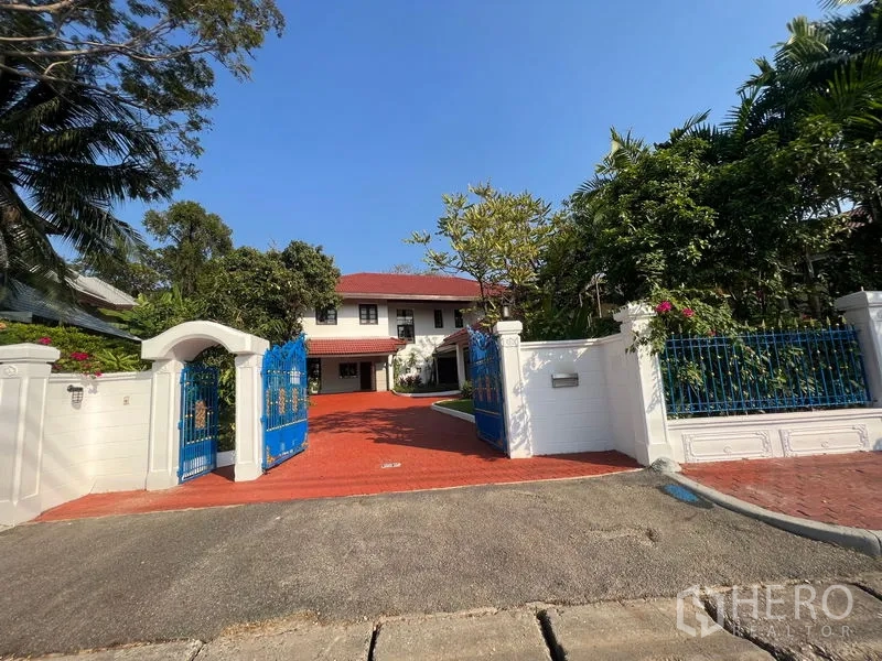 Detached House for rent in Bang Phli, Samut Prakan - Blue wrought-iron gate and white wall opening to a two-storey detached house with a red paved driveway and lush trees.