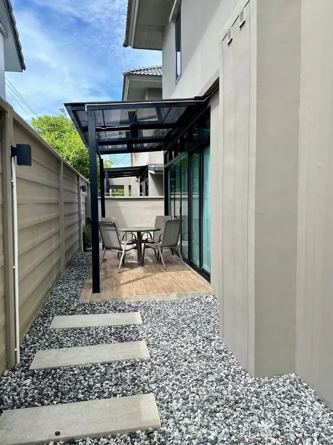 独立式住宅 出租在Samut Prakan - 侧院露台带玻璃顶棚、户外桌椅与碎石小径。