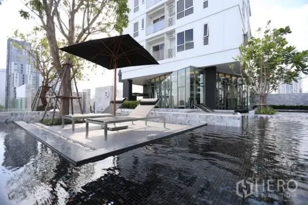 Condominium for rent or sale in Watthana, Bangkok - Resort‑style swimming pool with sun loungers and umbrella beside a modern glass‑walled condominium.