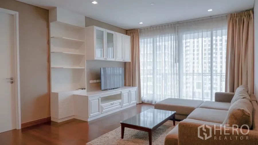 Condominium for rent in Watthana, Bangkok - Bright living room with L-shaped sofa, white built-in TV cabinet, and floor-to-ceiling windows with sheer curtains.