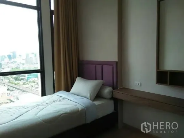 Condominium for rent in Huai Khwang, Bangkok - Second bedroom with single bed, built-in desk and floor-to-ceiling window with city view.