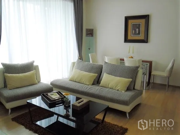 Condominium for rent or sale in Watthana, Bangkok - Bright living room with gray sofas, glass coffee table, dining nook, wood floors and sheer curtains in a Bangkok condo.