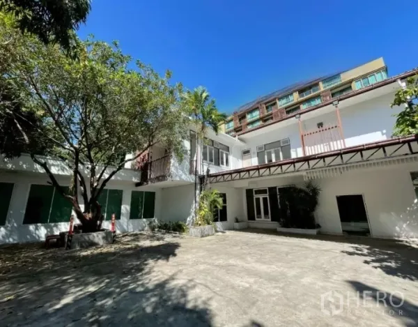 带办公室的家 出租在Watthana, Bangkok - 两层白色家庭办公楼，树荫庭院与宽阔停车区，蓝天下的外观。