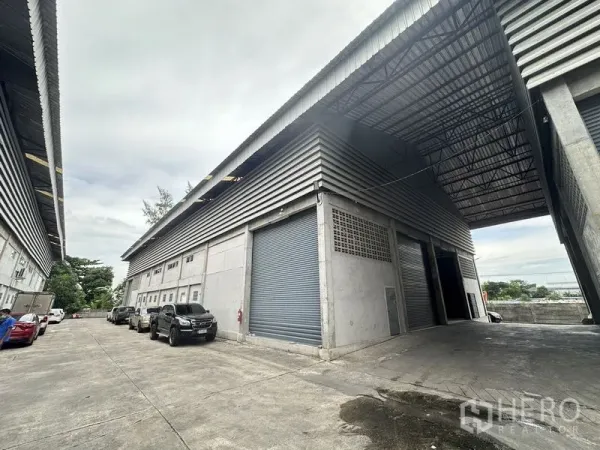 货栈 出租在Khlong Sam Wa, Bangkok - 大型仓库外观，金属板墙体、高挑屋顶与宽幅卷闸门，旁有车道与停车位