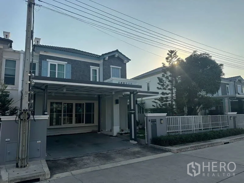 独立式住宅 出租在Prawet, Bangkok - 房屋正立面傍晚景，宽阔车道、金属大门与整洁绿化。