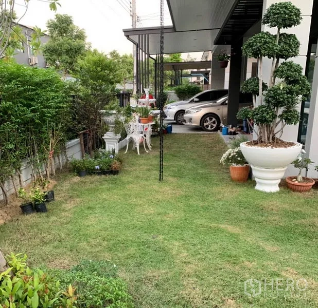 Detached House for rent or sale in Samut Prakan - Front garden with lawn, topiary and seating beneath a covered carport.