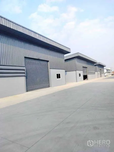 Warehouse for rent in Pathum Thani - Row of modern grey metal warehouses with roller shutter doors and a wide concrete driveway.