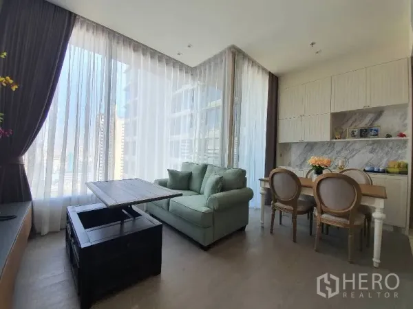 Condominium for rent or sale in Watthana, Bangkok - Bright living and dining area with floor-to-ceiling sheer curtains, city view, green sofa and built-in pantry with marble backsplash.