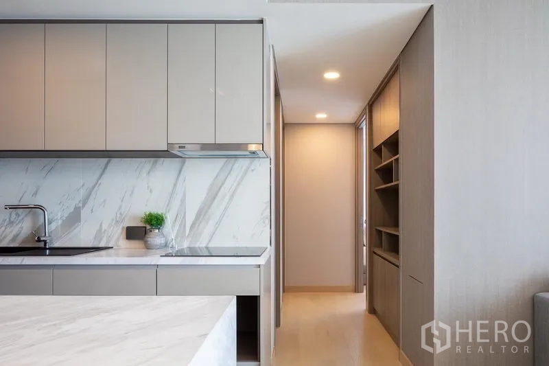 Condominium for rent in Khlong Toei, Bangkok - Close-up of sleek kitchen with marble backsplash leading to a hallway with custom shelving and warm lighting.