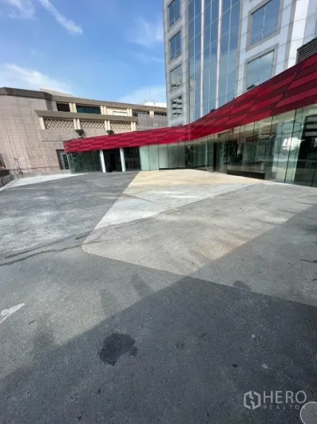 零售空间 出租在Pathum Wan, Bangkok - 现代零售门面，红色雨棚与宽阔广场相连，邻接玻璃幕墙建筑。