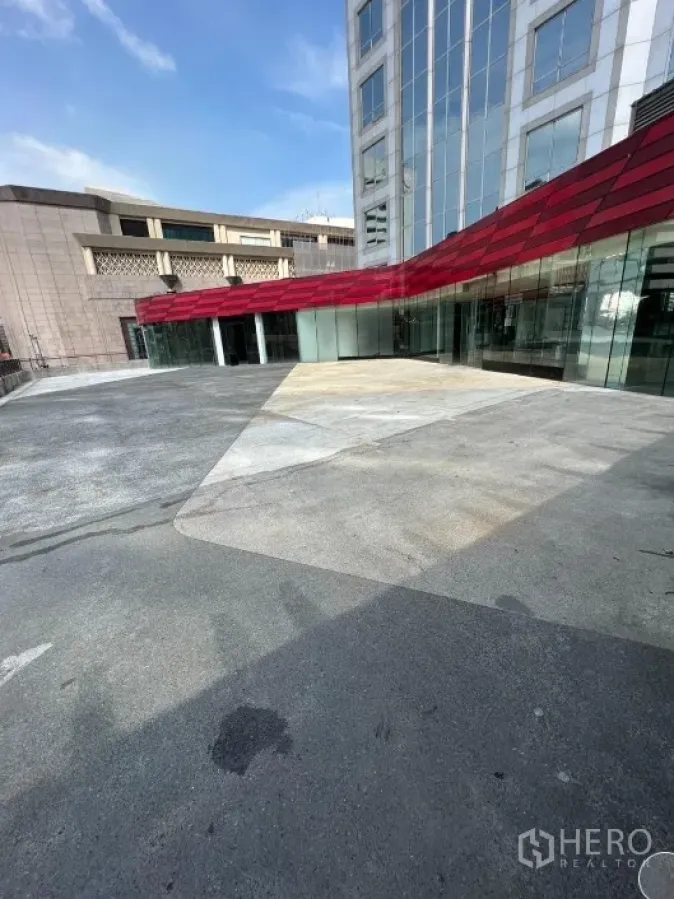 零售空间 出租在Pathum Wan, Bangkok - 现代零售门面，红色雨棚与宽阔广场相连，邻接玻璃幕墙建筑。