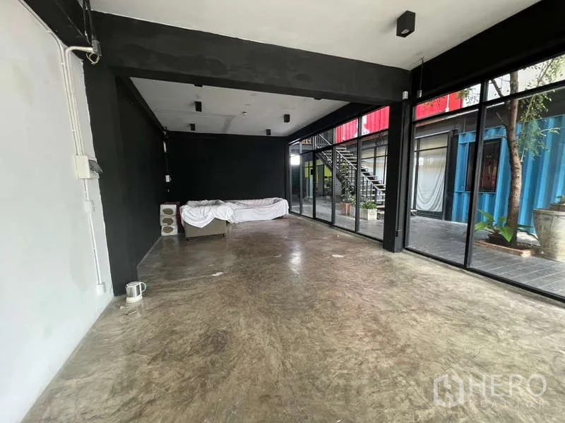 พื้นที่ขายปลีก ให้เช่า - มุมห้องกระจกสองด้าน ผนังสีดำ พื้นปูนเปลือย เห็นต้นไม้ภายนอก