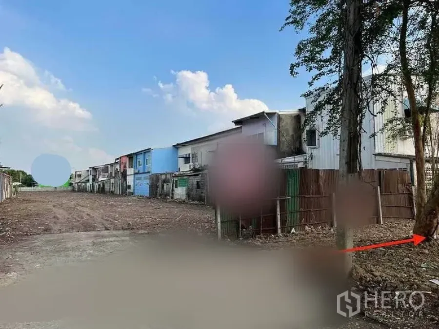 ที่ดิน ขายในMin Buri, Bangkok - มุมมองทางเข้าแปลงที่ดินกว้าง มีต้นไม้และรั้วข้างทาง อาคารพาณิชย์เรียงตลอดแนว