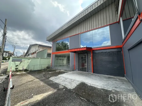 Warehouse for rent in Suan Luang, Bangkok - Angled exterior showing gated forecourt, glass storefront and black roller shutter entry.