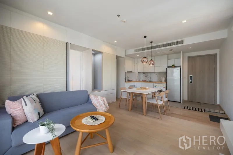 Condominium for rent in Watthana, Bangkok - Living area with sofa, round coffee tables and dining set next to a white kitchen.