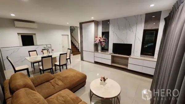 Detached House for rent in Prawet, Bangkok - Open-plan living and dining area with sectional sofa, TV wall and staircase.