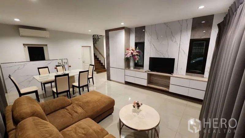 Detached House for rent in Prawet, Bangkok - Open-plan living and dining area with sectional sofa, TV wall and staircase.