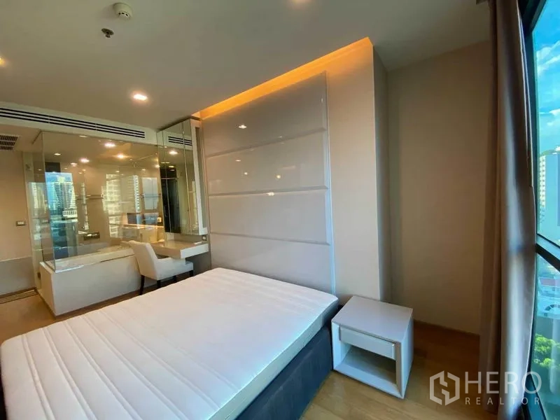 公寓 出租在Bang Rak, Bangkok - 明亮卧室，玻璃隔断与嵌入式床头和书桌，窗外城市景观。