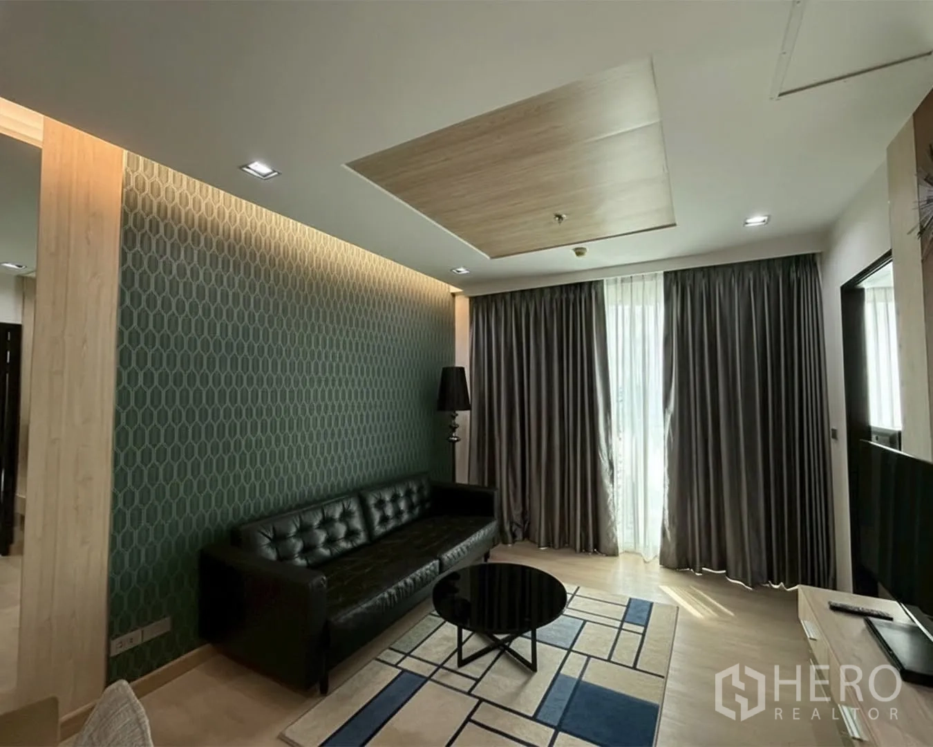 Condominium for rent or sale in Watthana, Bangkok - Modern living room with leather sofa, patterned accent wall, coffee table, and full-length curtains.