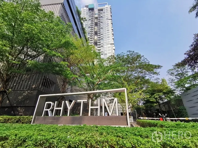 Condominium for rent in Khlong Toei, Bangkok - Condominium tower exterior with RHYTHM sign and landscaped entrance surrounded by trees.