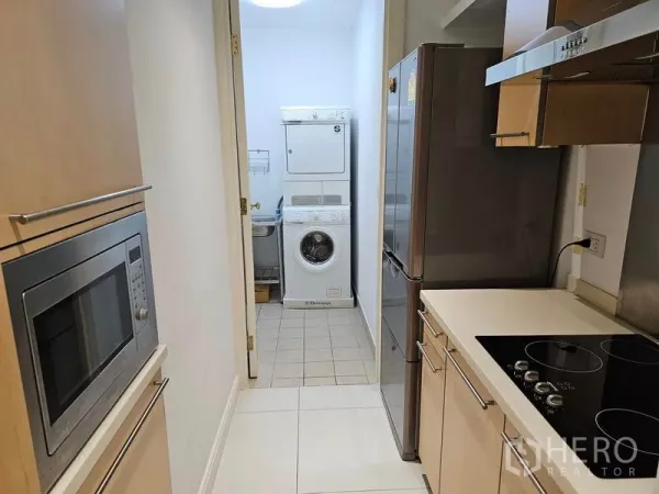Condominium for rent or sale in Pathum Wan, Bangkok - Galley kitchen with built-in appliances leading to a separate laundry room with stacked washer and dryer.
