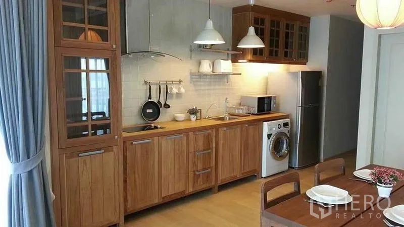 Condominium for rent or sale in Watthana, Bangkok - Open kitchen with wooden cabinetry, subway tile backsplash, fridge and washing machine beside dining table.