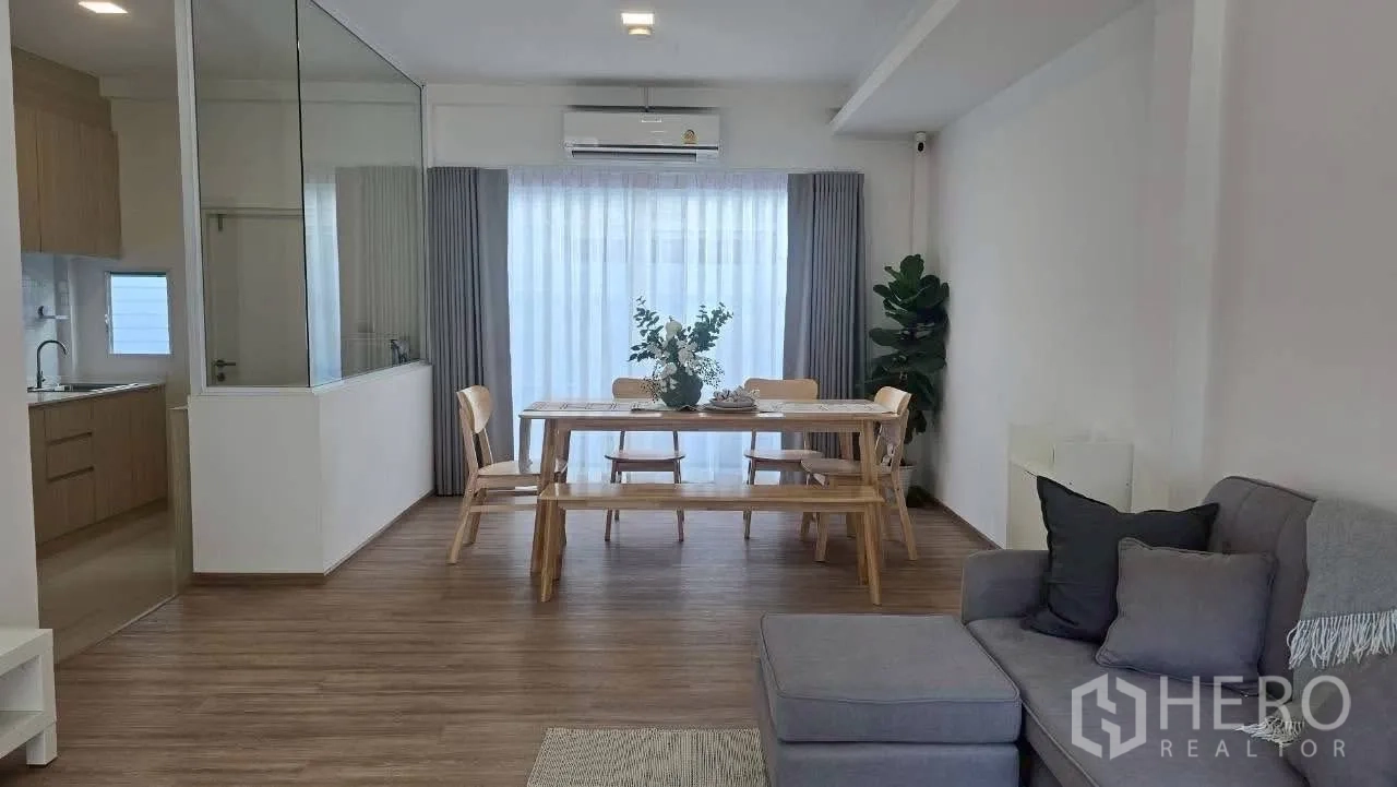 Townhouse for rent in Prawet, Bangkok - Open-plan living and dining room with glass-partitioned kitchen and full-height curtains.