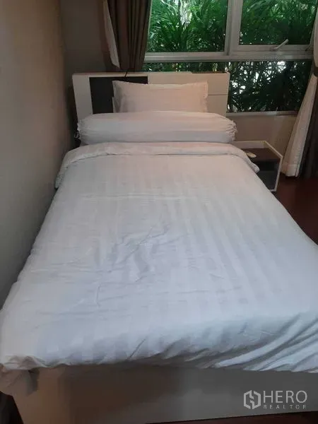 Condominium for rent or sale in Huai Khwang, Bangkok - Single bedroom with white bedding and a large window overlooking garden plants.