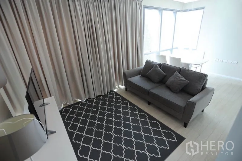 Condominium for rent or sale in Watthana, Bangkok - Cozy living room with grey sofa, patterned rug, TV console, and full-length curtains.