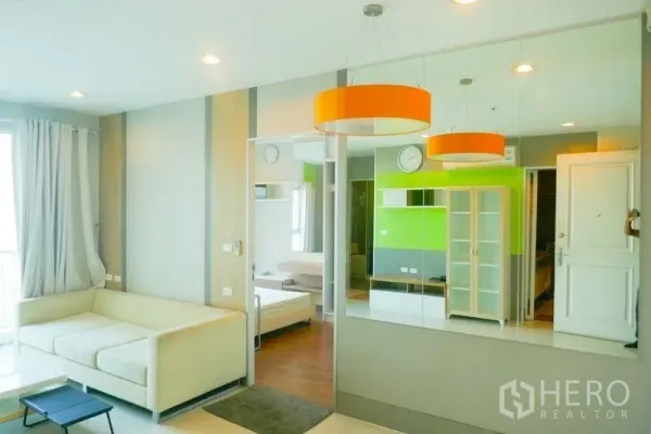 Condominium for rent or sale in Khlong San, Bangkok - Open-plan lounge with orange pendant lights, mirror wall and sofa, view into bedroom.