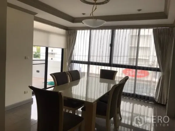 Condominium for rent in Watthana, Bangkok - Dining area with a six‑seat table beside floor‑to‑ceiling windows and sheer curtains, with balcony access visible.