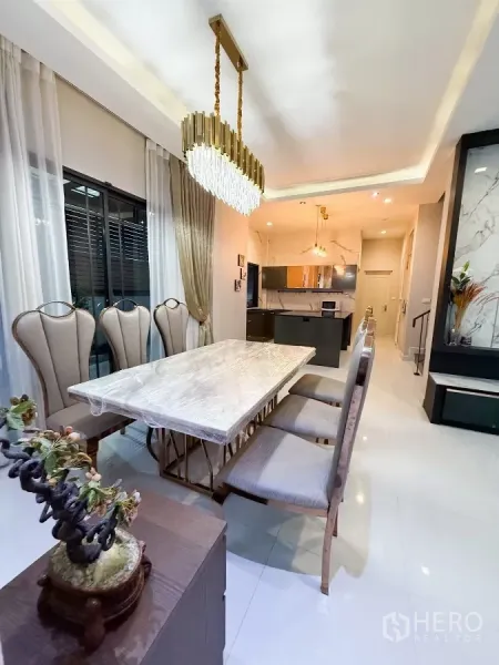 Detached House for rent or sale in Bang Phli, Samut Prakan - Dining area with six upholstered chairs and a marble‑top table beside windows, looking toward a modern kitchen island.