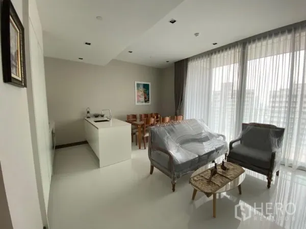 Condominium for rent in Khlong Toei, Bangkok - Open-plan lounge, dining table and kitchen island beside wide balcony-style windows for abundant light.