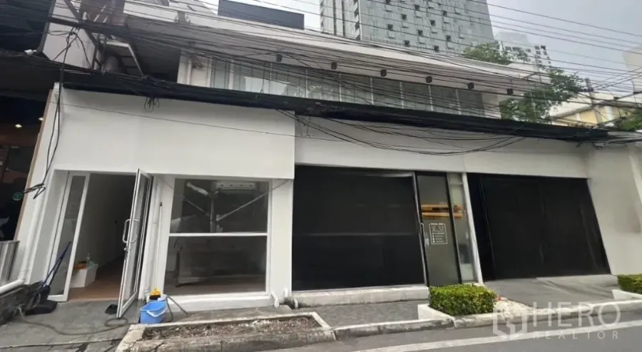 零售空间 出租在Watthana, Bangkok - 通罗街边店面外观，白色立面，宽幅玻璃与卷帘，低层建筑前有高楼背景。