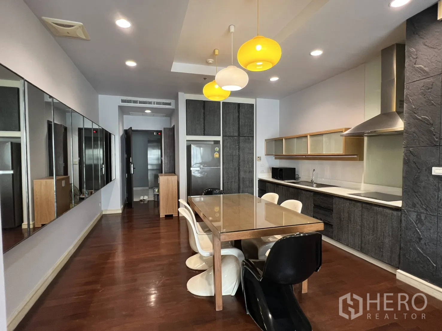 Condominium for rent in Watthana, Bangkok - Open-plan kitchen and dining area with wood floors, long table, pendant lights, sleek cabinetry, and a stainless range hood.