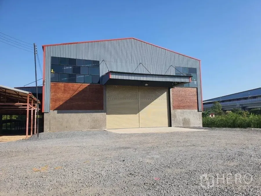 货栈 出租在Mueang Pathum Thani, Pathum Thani - 金属外墙仓库外观，宽幅卷帘门、山墙屋顶与宽阔碎石前场