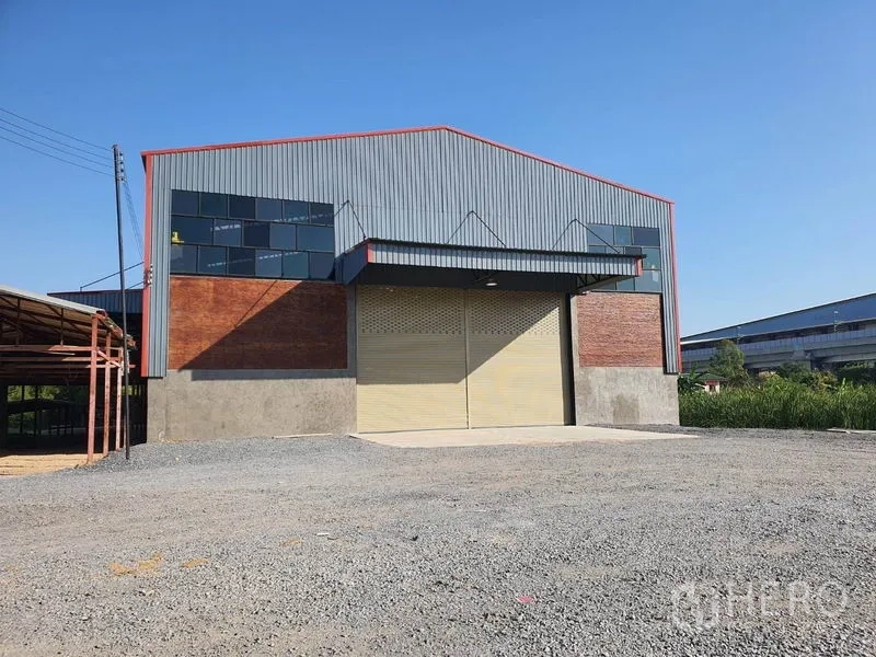 货栈 出租在Mueang Pathum Thani, Pathum Thani - 金属外墙仓库外观，宽幅卷帘门、山墙屋顶与宽阔碎石前场