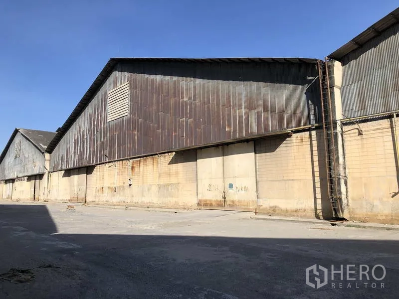 Warehouse for rent in Phra Nakhon Si Ayutthaya - Side exterior of long corrugated metal warehouse with concrete base and driveway under blue sky.