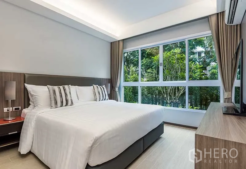 Apartment for rent in Huai Khwang, Bangkok - Master bedroom with king bed, large windows, and leafy garden outlook.