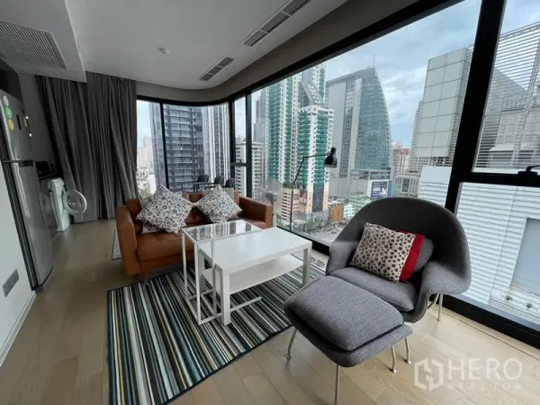 Condominium for rent or sale in Watthana, Bangkok - Corner lounge with wraparound glass walls, city skyline views, sofa, armchair and coffee tables on a striped rug.