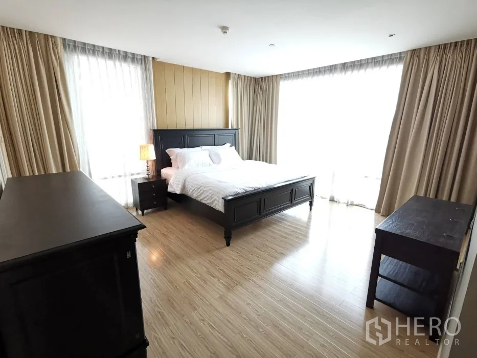 Condominium for rent in Watthana, Bangkok - Sunlit master bedroom with dark wood bed and furniture, full-height curtains, and warm wood flooring.