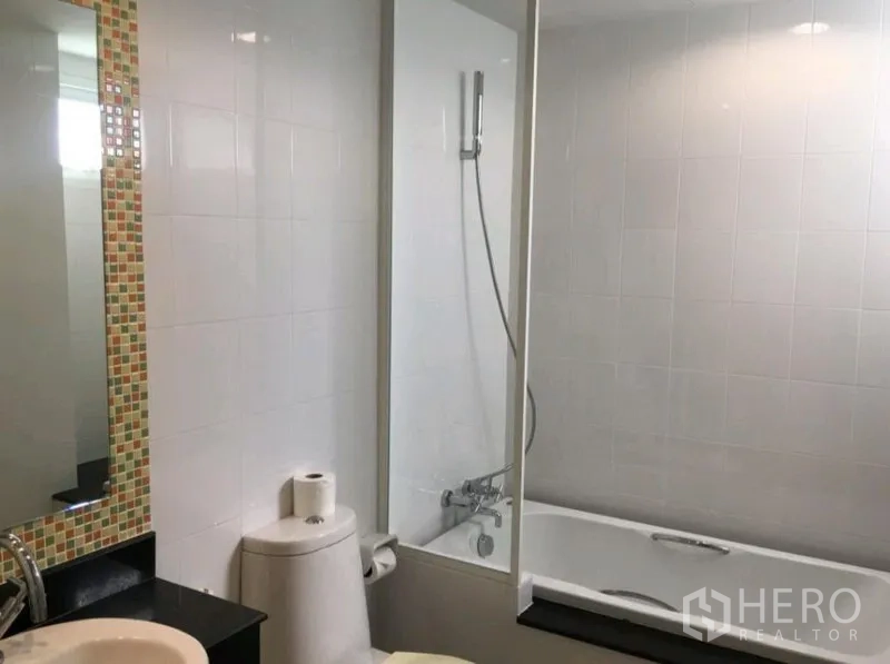 Condominium for rent in Phra Khanong, Bangkok - Modern bathroom with bathtub and glass shower screen, handheld shower, toilet and a large mirror with mosaic trim.