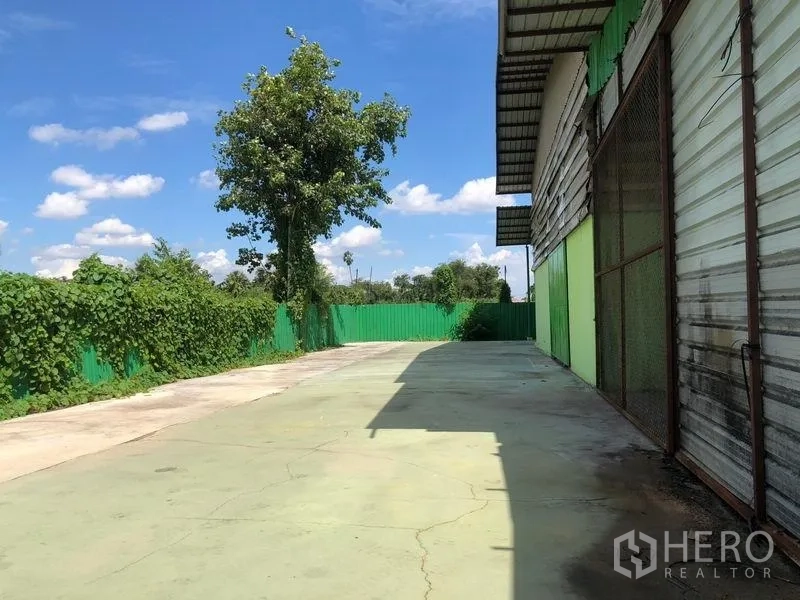 Factory for rent in Sam Khok, Pathum Thani - Sunny side yard with perimeter fence and access lane beside the warehouse facade.