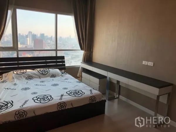 Condominium for rent or sale in Huai Khwang, Bangkok - Bright bedroom with a large window, skyline view, bed and a long built-in desk.