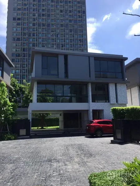 独立式住宅 出租在Watthana, Bangkok - 现代三层独栋，落地窗与封闭车道，前停红色车辆，背景为高层建筑。