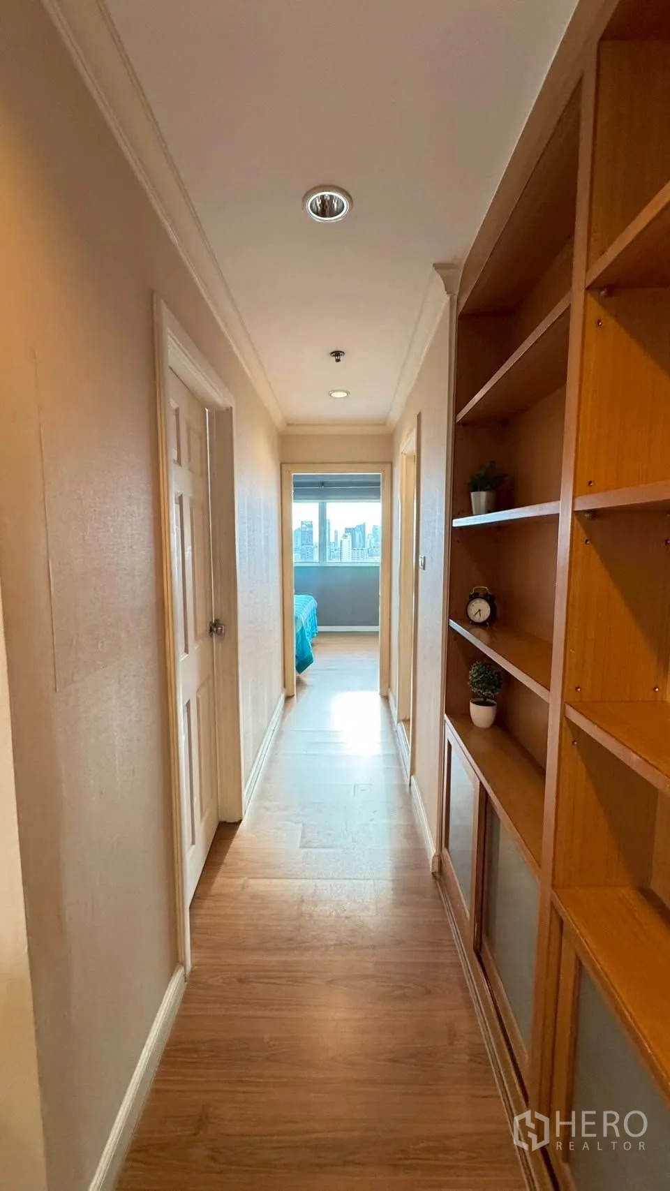 Condominium for rent or sale in Watthana, Bangkok - Hallway with wooden flooring and shelving, opening to bedrooms with skyline view at the end.