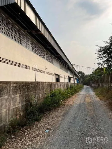 货栈 出售在Bang Bua Thong, Nonthaburi - 仓库沿碎石通道的一侧立面，旁有围墙与绿化带。