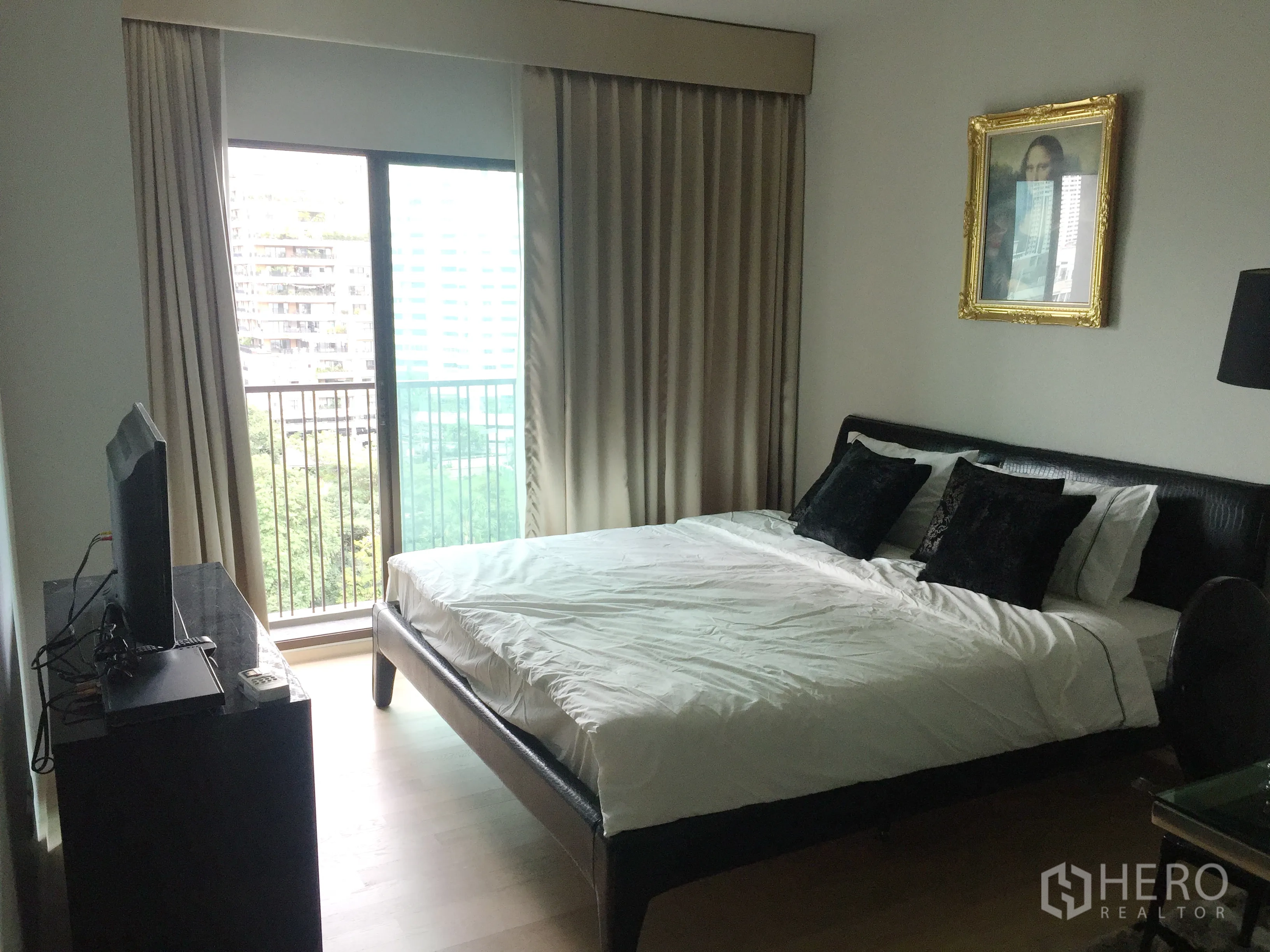 คอนโด ให้เช่าในBangkok - ห้องนอนเตียงขนาดใหญ่ ม่านสูง ตู้ทีวี และประตูกระจกออกสู่ระเบียงมองเห็นวิวเมืองและต้นไม้